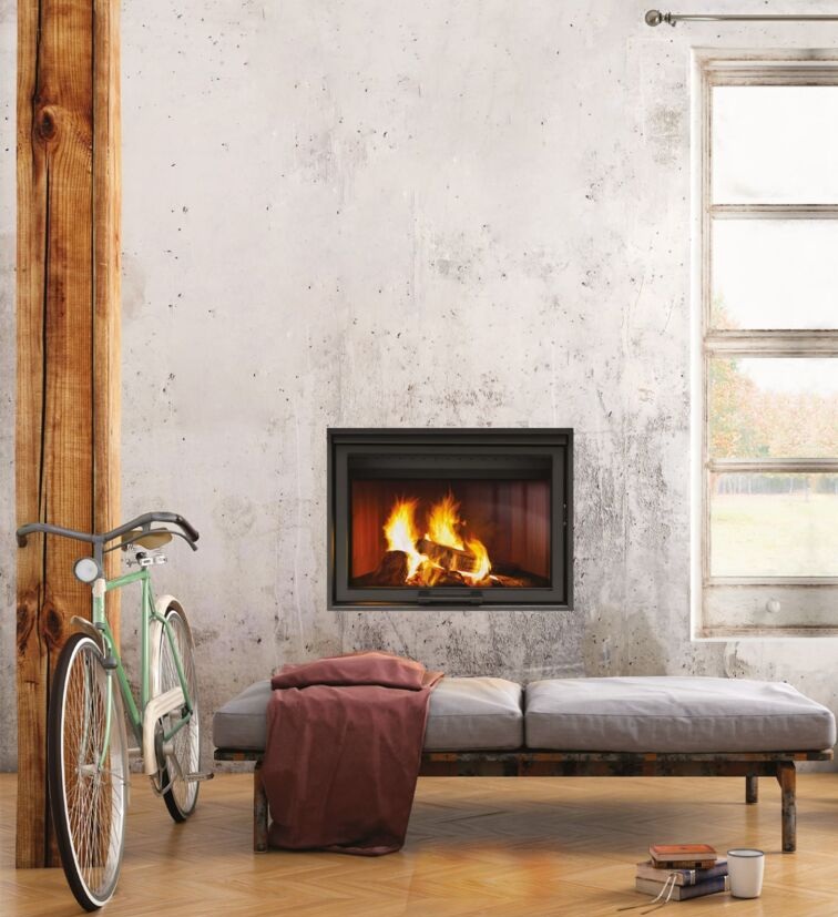 Camino a legna incasso acceso su parete di cemento grezzo, con daybed grigio, plaid bordeaux e bici verde su parquet